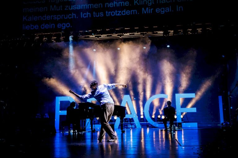 Ein Tänzer, der vor dem Schriftzug "Peace" auf der Bühne tanzt in blauem Licht